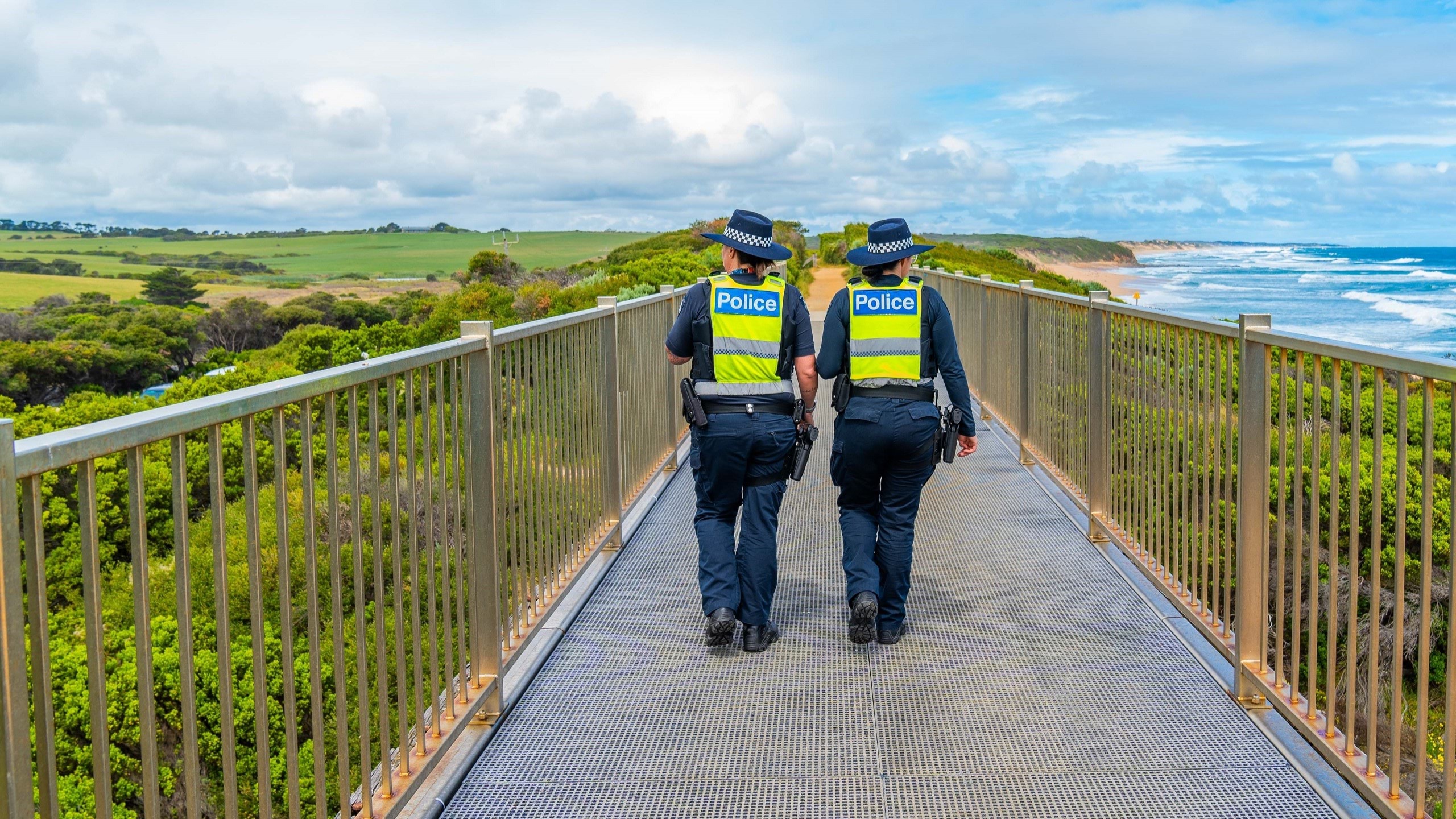 Two police officers walking along a jetty with green brushland to their left and the ocean plus sand dunes to their right