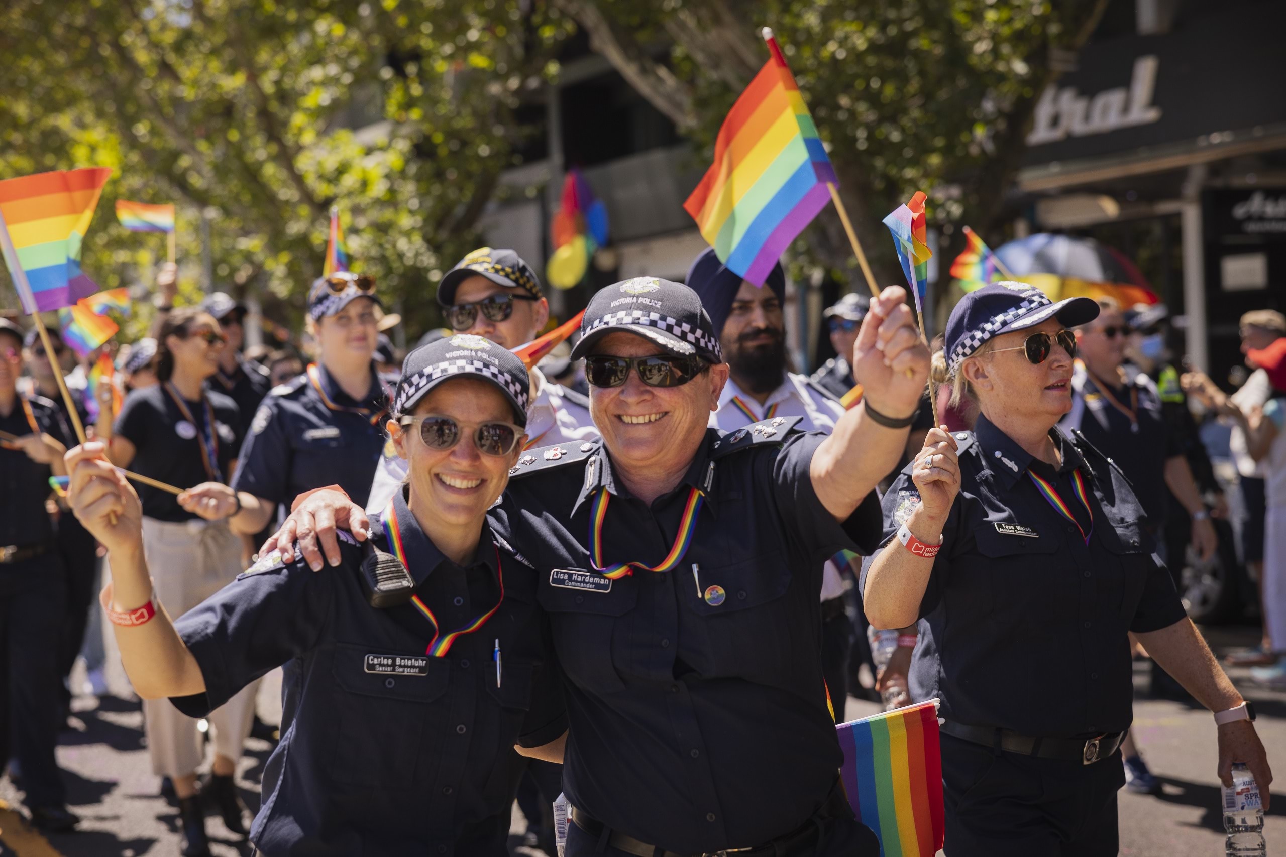 Police officers walking along the road and footpath during the day in front of shops taking part in a peaceful pride march for the LGBTQIA+ community. Two police officers in uniform are smiling at the camera waving rainbow-coloured flags waving 