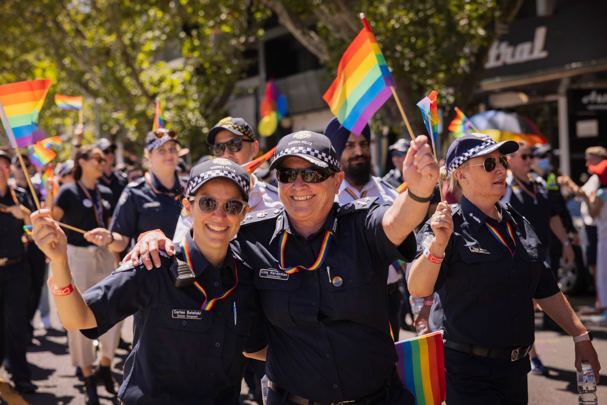 Supporting family and friends Police officers walking along the road and footpath during the day in front of shops taking part in a peaceful pride march for the LGBTQIA+ community. Two police officers in uniform are smiling at the camera waving rainbow-coloured flags waving