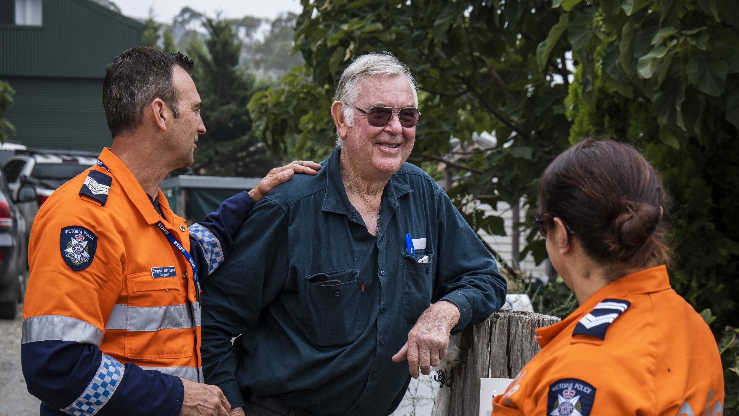 Two country-based police officers dressed in bright orange emergency services colour standing engaged in smiling conversation with an older local male as part of friendly community policing