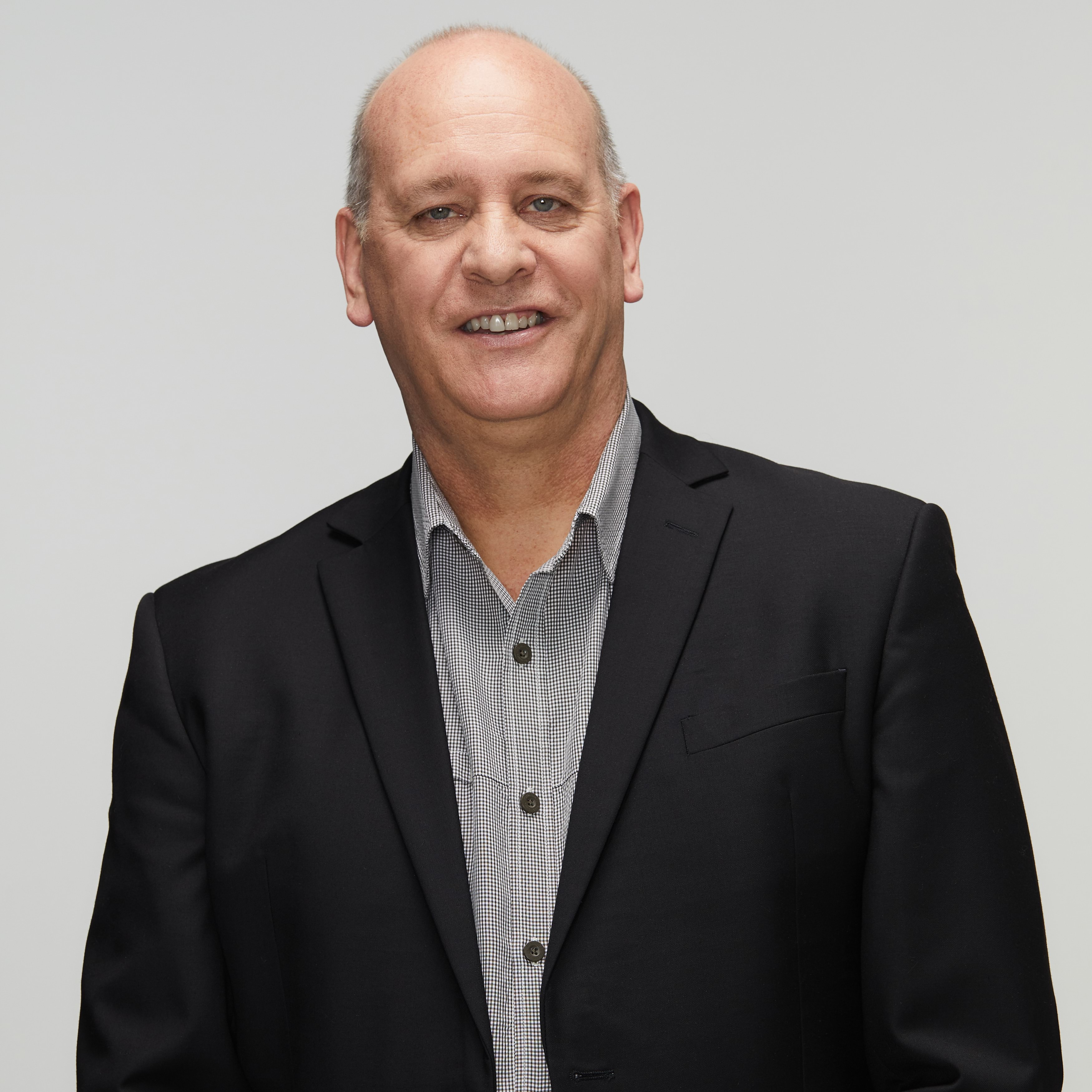 Image of current police chaplain smiling at the camera wearing a grey and white pattern shirt with a navy suit jacket on top. Late forties, male with light skin and light, receding hair.