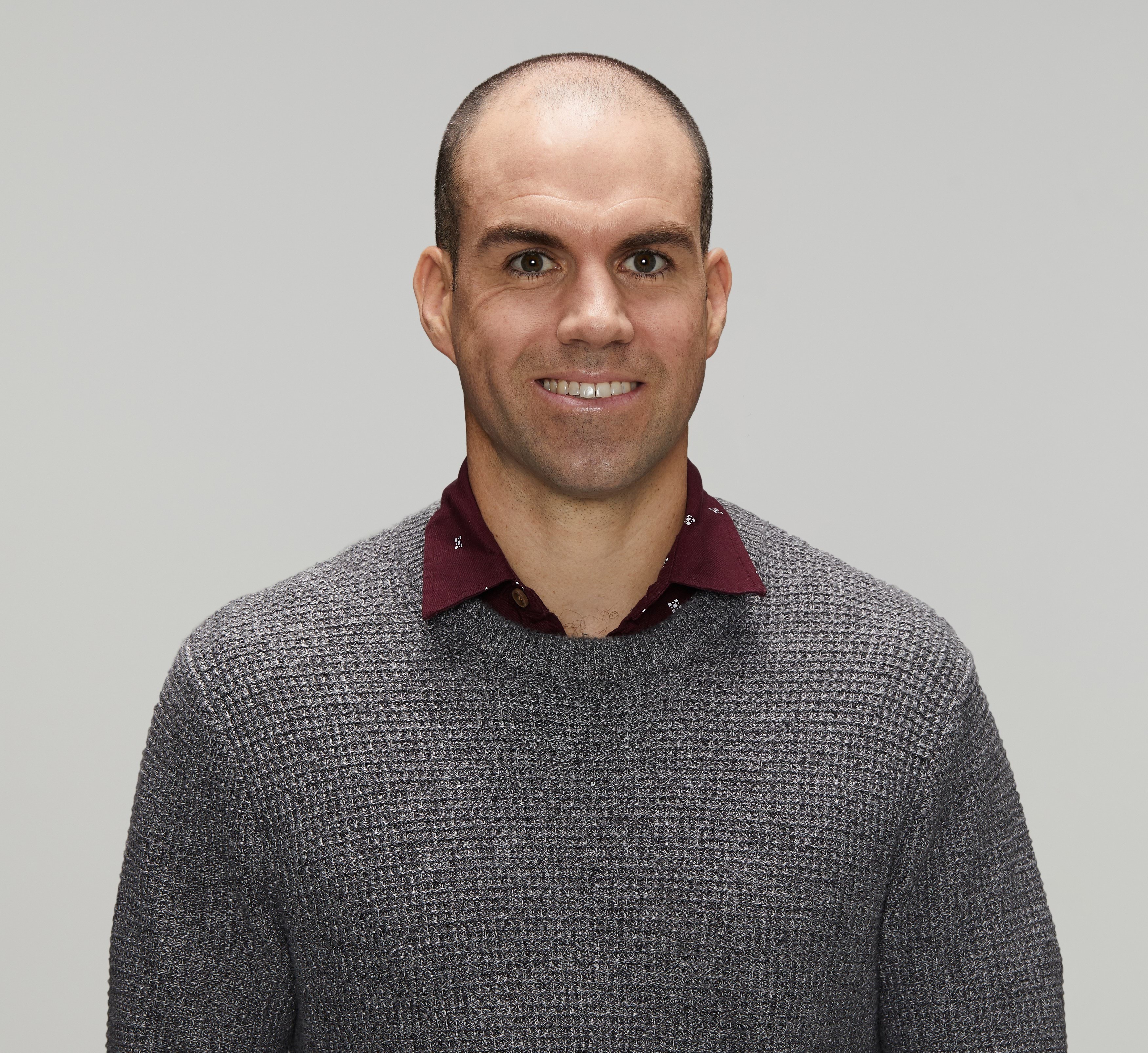 Police officer in civilian clothing smiling at the camera. White male, early forties with dark receding hair, wearing a maroon collared shirt with a smart casual metal grey jumper over the top.