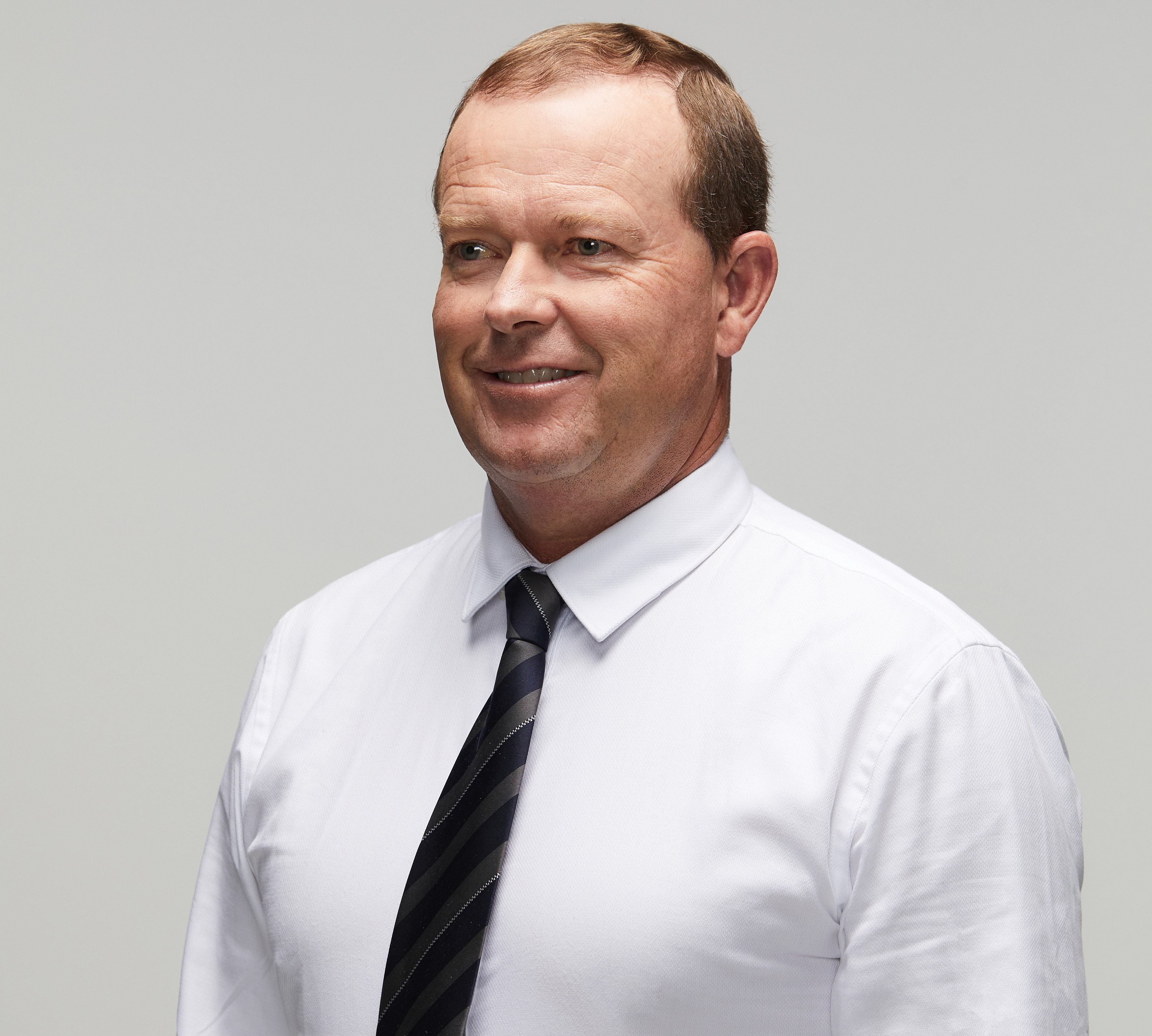 Police officer in civilian clothing smiling to left of camera, wearing long-sleeved white shirt with black and silver stripped tie; white male, mid to late forties with thinning nutbrown, coloured hair