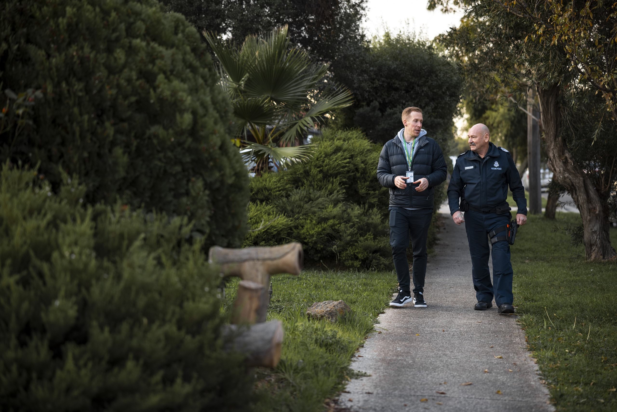 Social worker and Senior Sergeant police officer talking and walking together along a tree-lined footpath. The social worker is wearing a staff identification lanyard. The police officer is wearing standard issue navy blue uniform, long pants and long-sleeved jacket, with a name badge on one side of his chest and Victoria Police silver, star logo with Victoria Police written underneath on the other side of his chest.