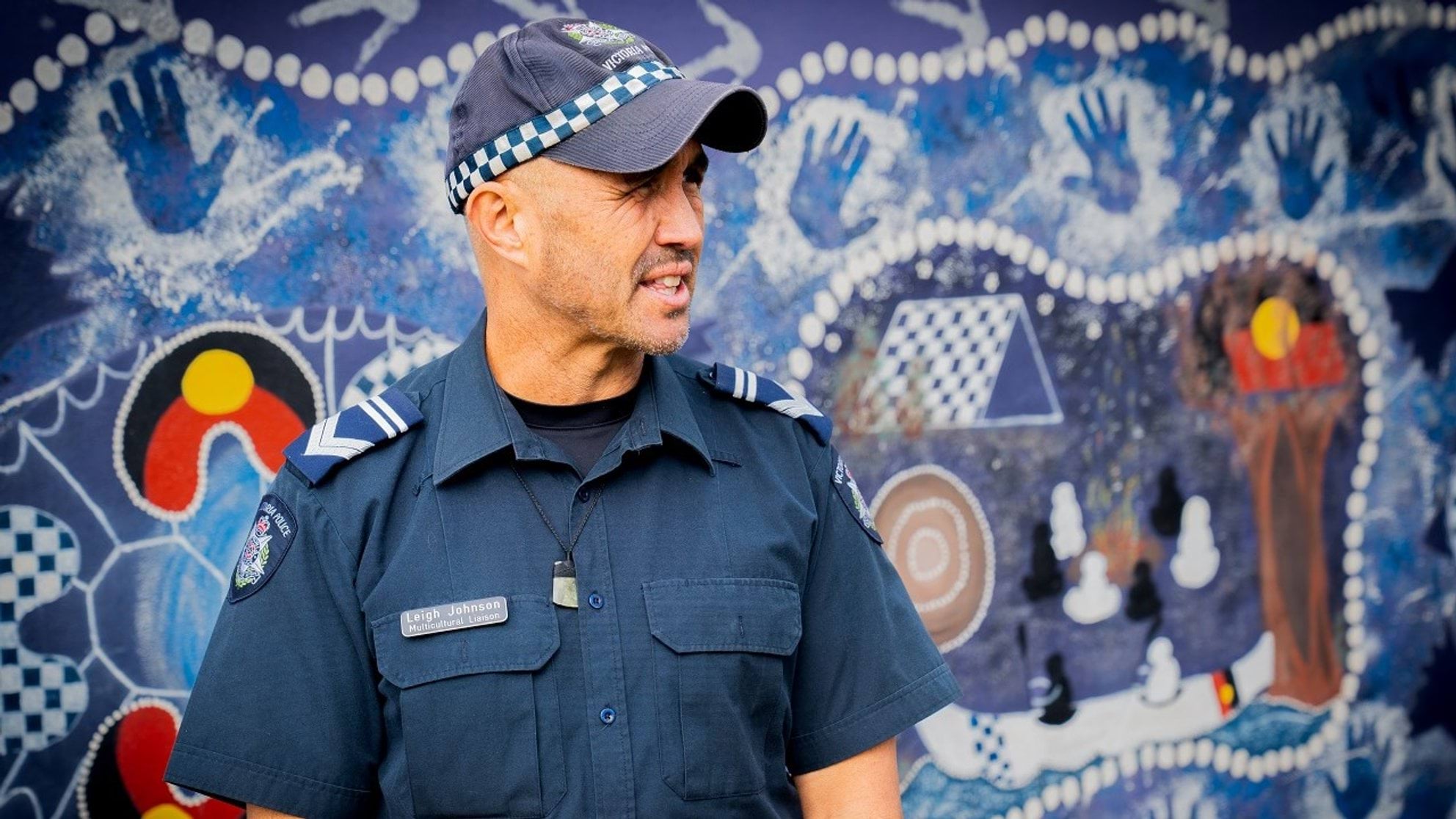 Wellbeing support for employees and their families Uniformed police officer looking off to the side standing in front of colourful mural of painted Indigenous Aboriginal artwork. Police officer is middle-aged man with olive skin wearing standard issued navy blue police uniform. Blue short-sleeved, collared shirt with embroidered Victoria Police badge on right shoulder, and baseball-style peeked cap with Victoria Police written across top front. Police officer also wears a badge pinned on right side of their chest that reads Multicultural Liaison Officer.