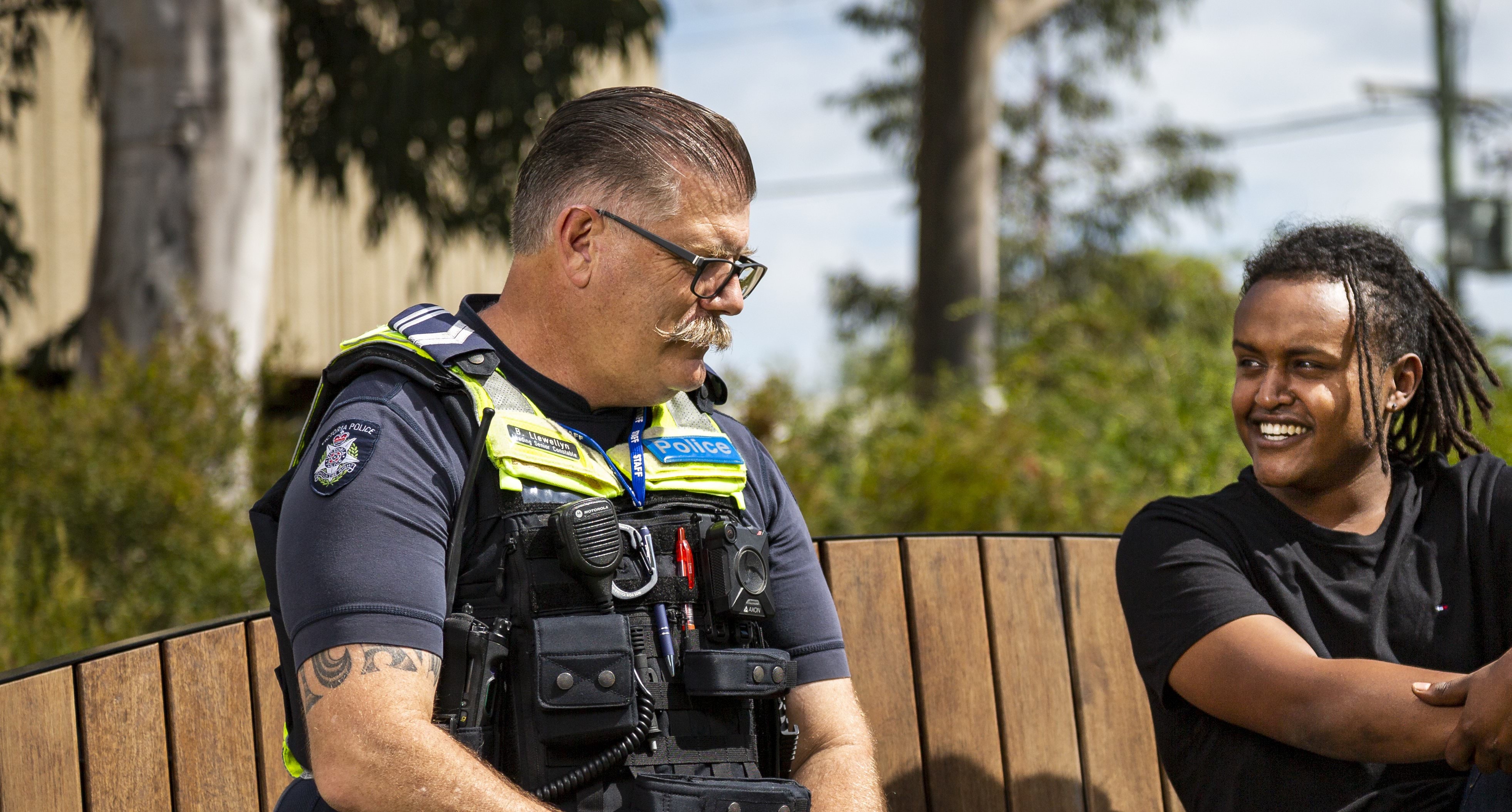 Two people sitting on a beach, talking, smiling and listening to one another; one is a male uniformed police officer, the other is a public citizen with shoulder-length braided Cornrows and a black T-shirt.