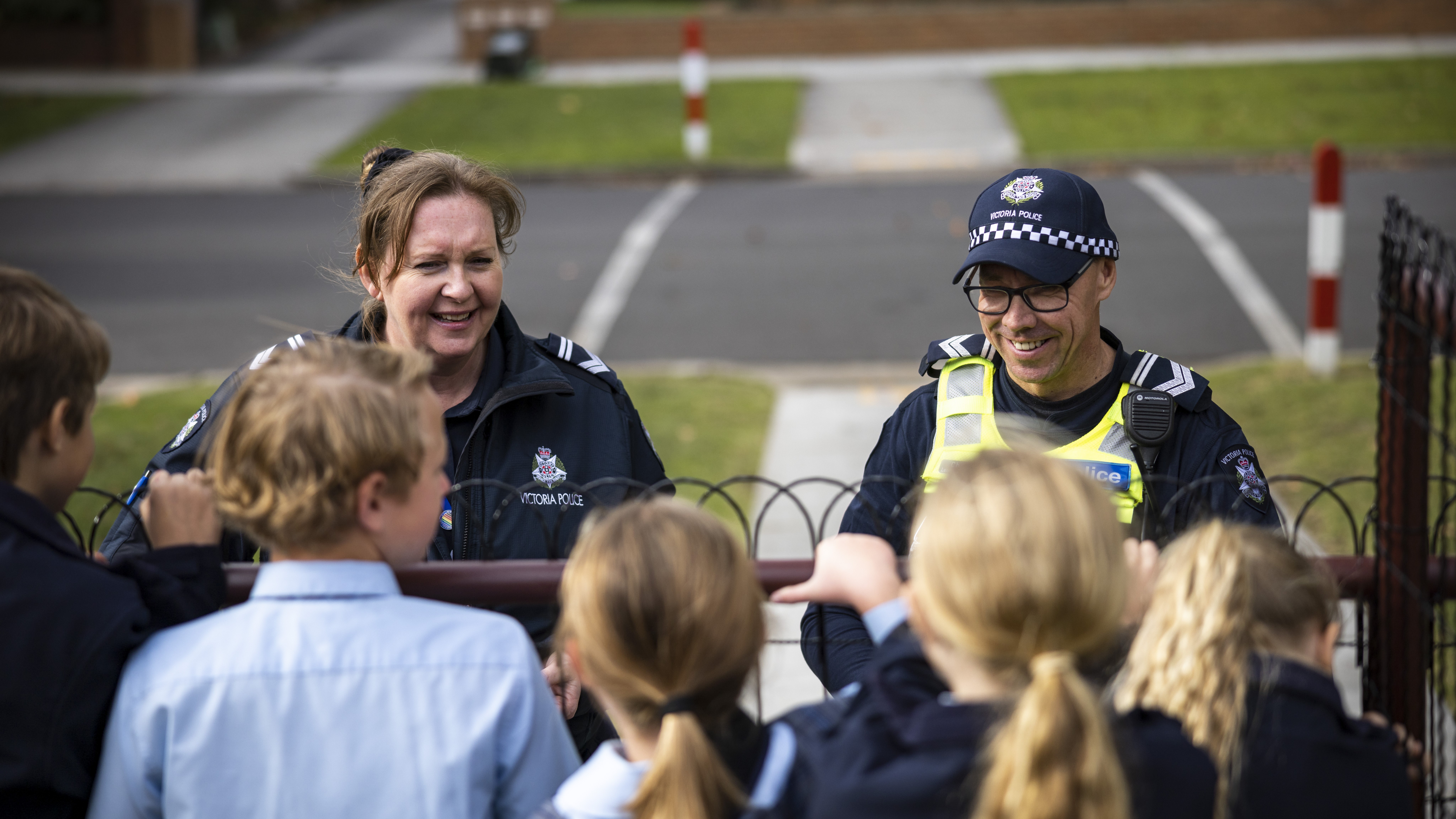 Two uniformed police officers engaging a local primary school children in friendly conversation in the local community. Wearing standard issue navy blue and white police uniform, Victoria Police is written across the top front of the baseball style peeked cap one of the officers is wearing. The Victoria police silver star logo with Victoria Police written underneath is on the front left chest pocket of the other police officer.