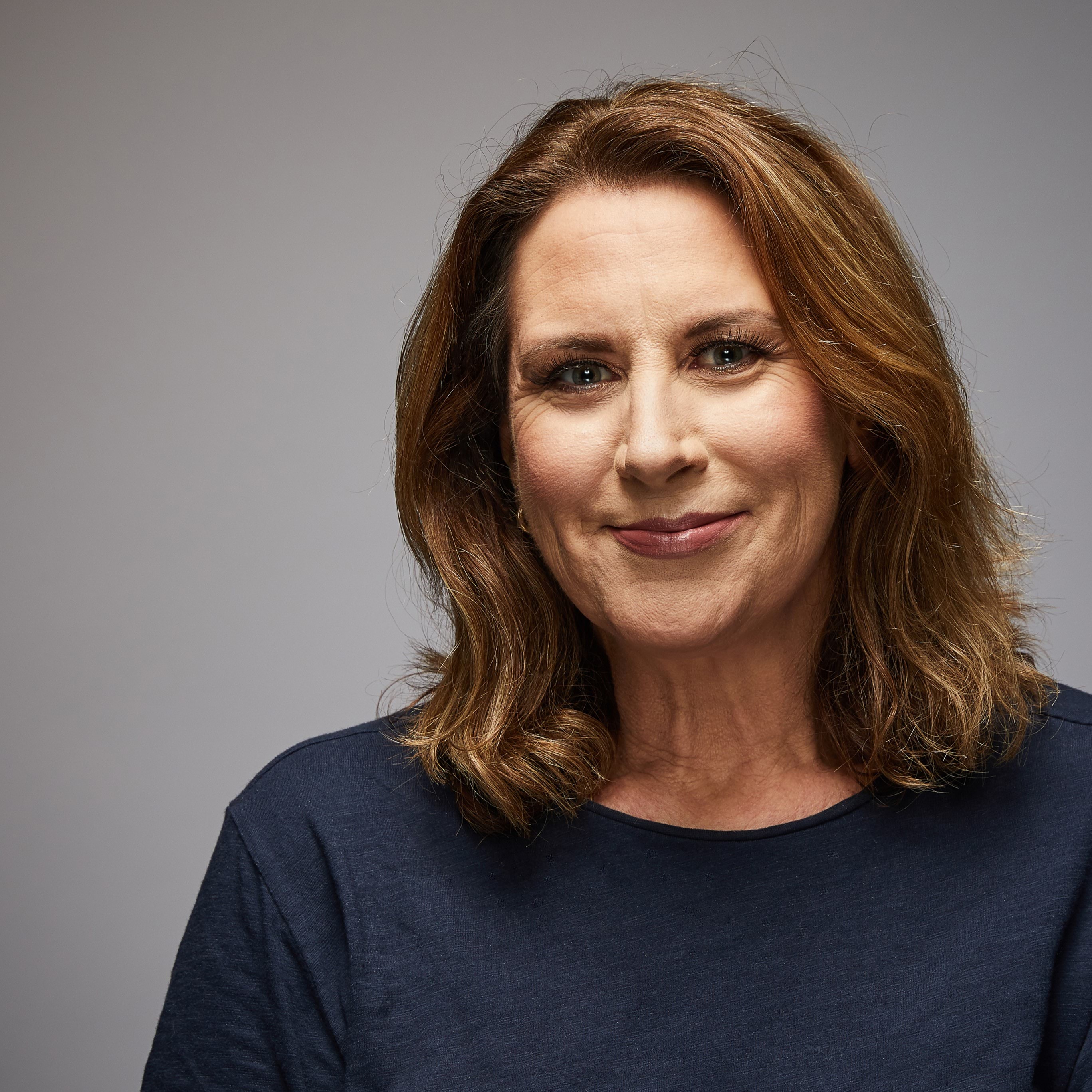 Female police officer in civilian clothing smiling at camera, wearing long-sleeved smart casual navy blue top; Caucasian female, mid to late forties with shoulder-length honey brown, coloured hair